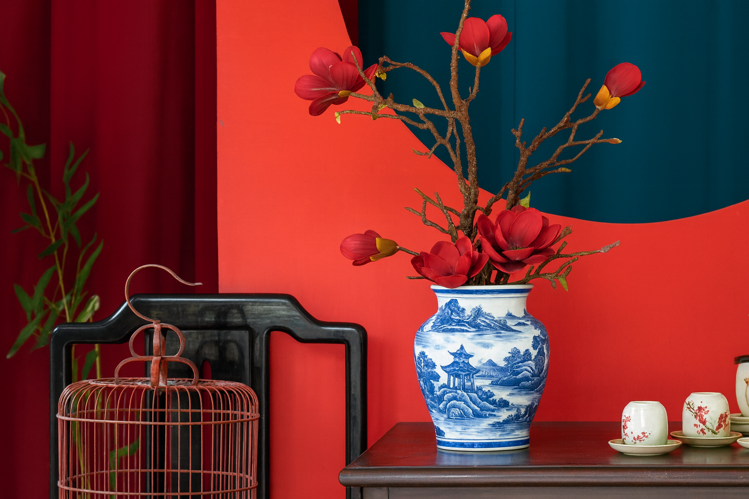 Blue and white Canton porcelain vase with a traditional landscape design, filled with red magnolia flowers, sitting on a wooden table beside teacups in a vibrant red and teal interior.