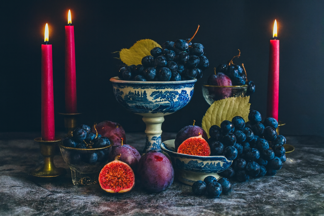 Still life of figs and grapes with fig-colored taper candles in brass holders and an antique blue and white transferware compote, styled on a dark textured surface.